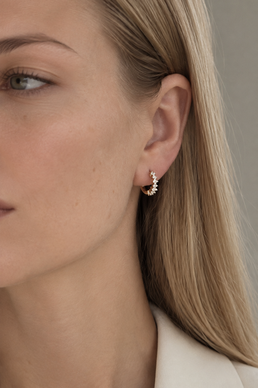 A close-up of a woman's ear wearing a small gold crescent-shaped huggie hoop earring with tiny clear stones, shown against a soft gray background.