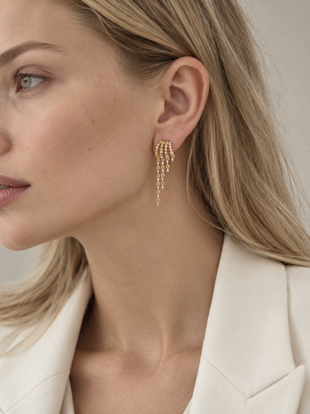 A close-up of a woman wearing a delicate gold dangle earring with multiple cascading chains of small beads, paired with a cream-colored blazer.