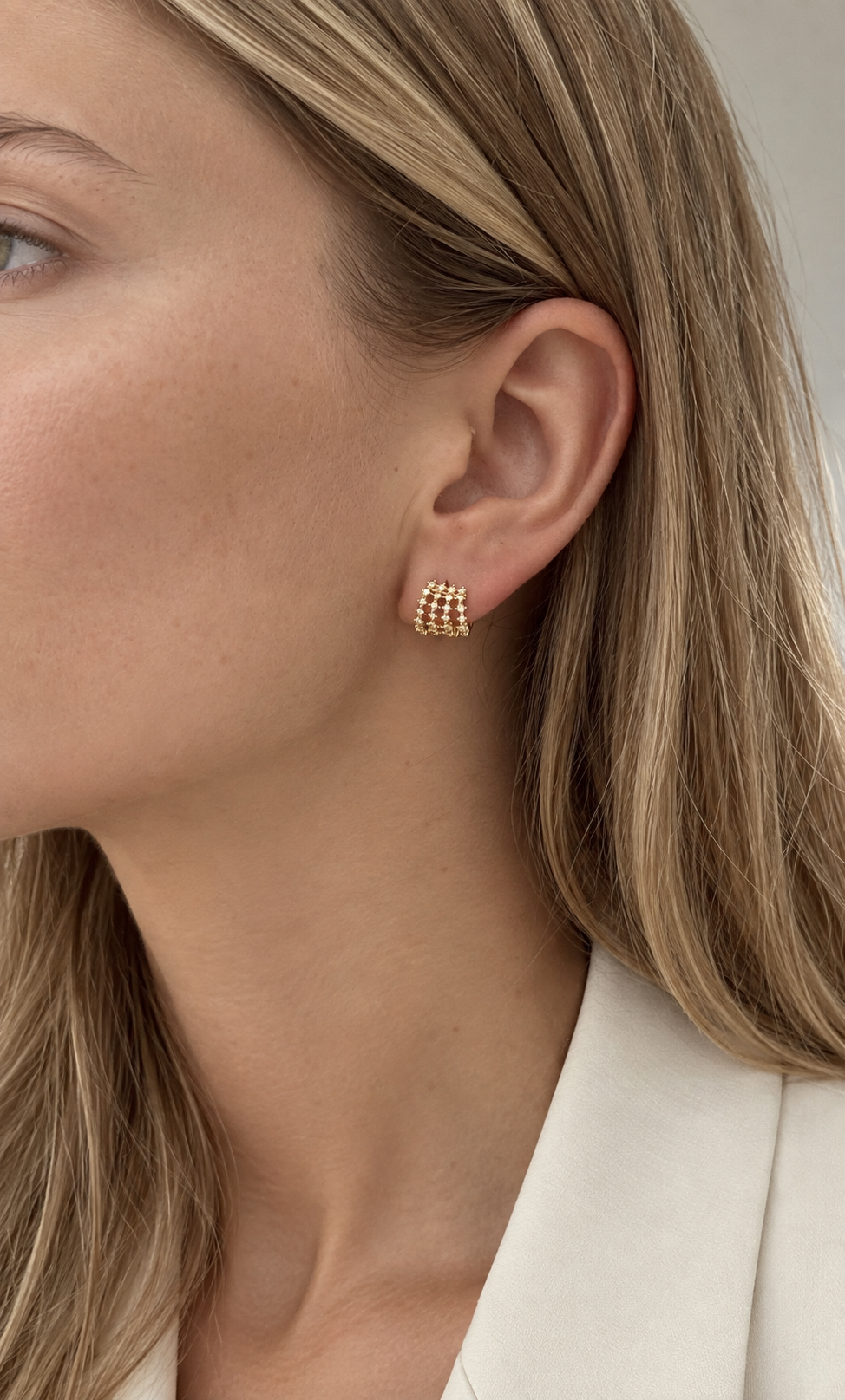 A close-up of a woman's ear wearing a small gold stud earring with a textured, basket-like grid pattern.